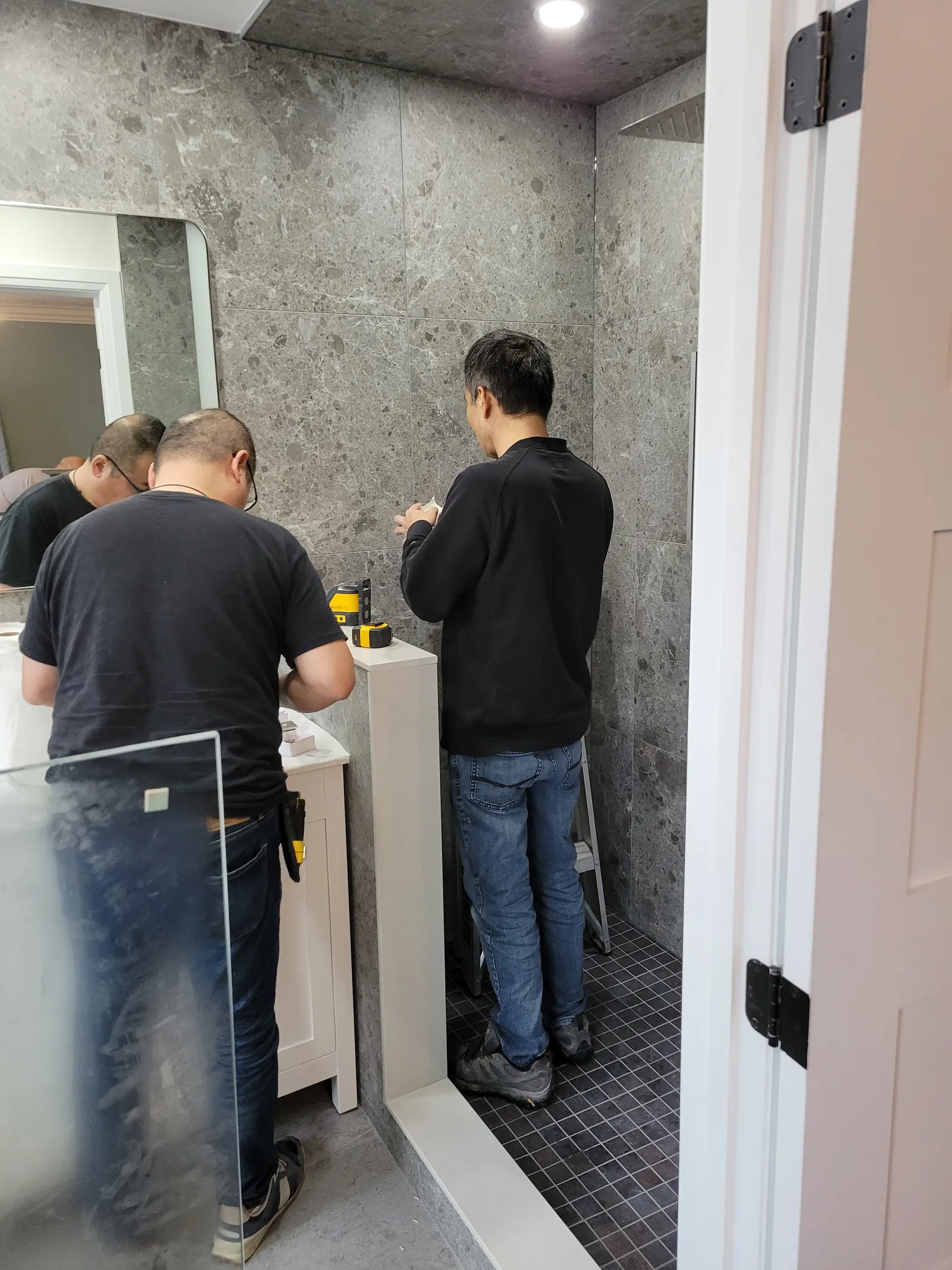 Frameless glass shower enclosure being fitted over the dark mosaic shower floor