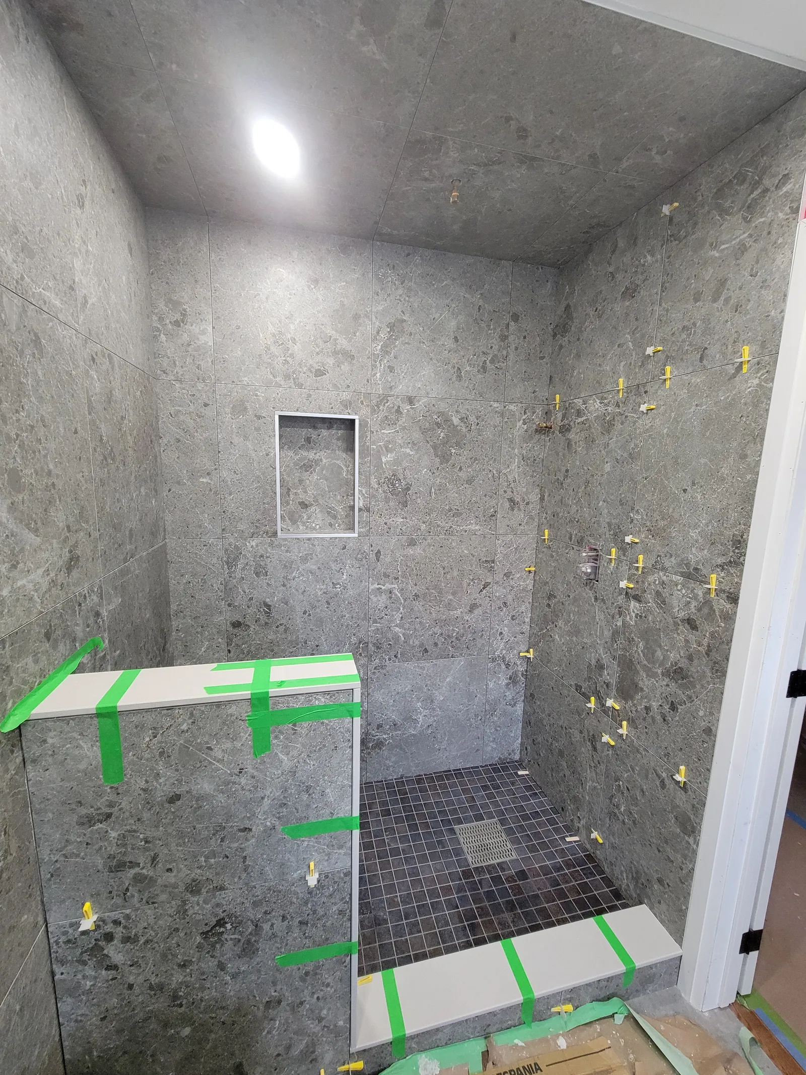 Large-format grey porcelain shower tile with a recessed niche and mosaic floor, taped during installation