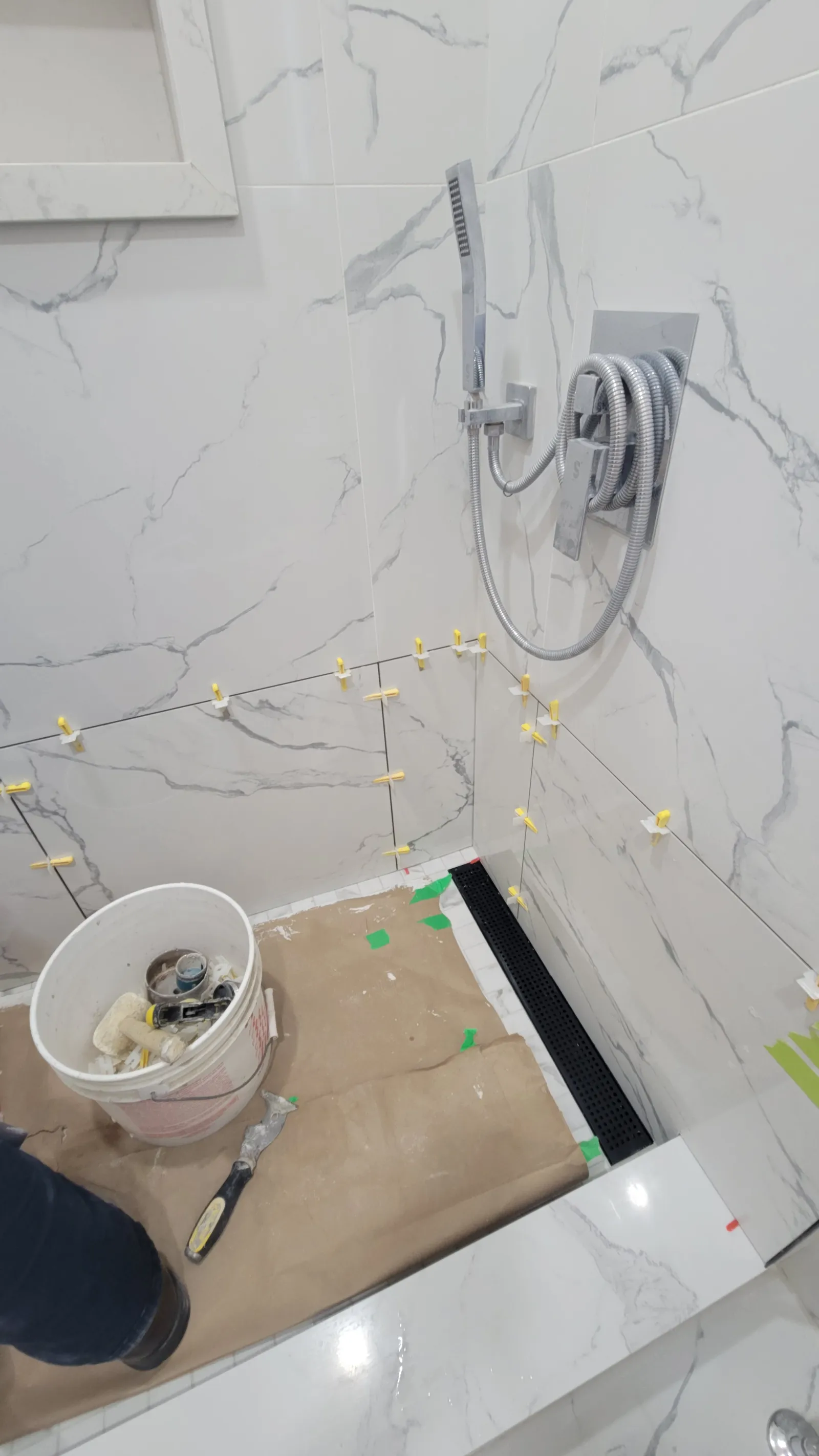 Re-tiled shower walls in Newmarket with levelling clips still in place, the valve and hand shower reinstalled, and the new floor protected with paper